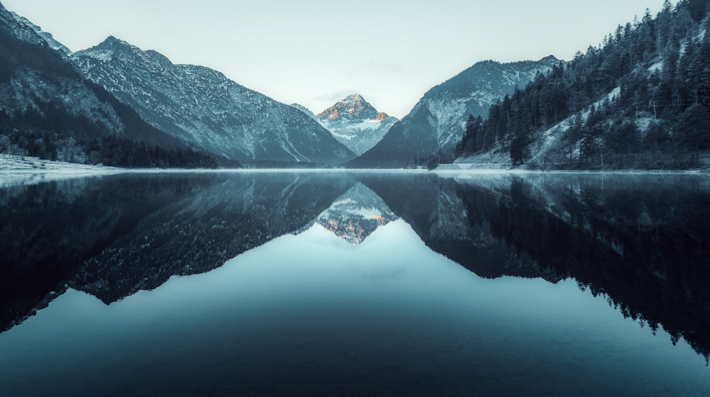 Berglandschaft mit spiegelglattem See und schneebedeckten Gipfeln im Hintergrund.