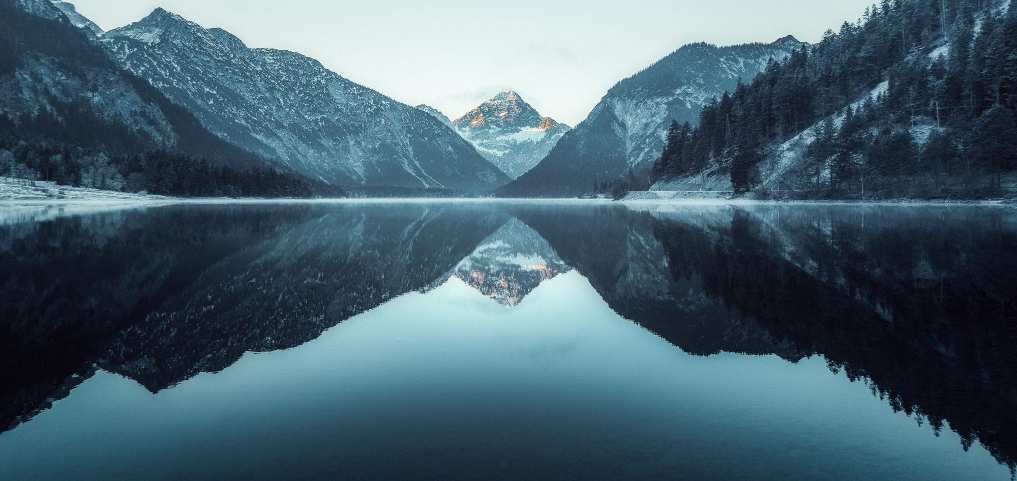 Berglandschaft mit spiegelglattem See und schneebedeckten Gipfeln im Hintergrund.